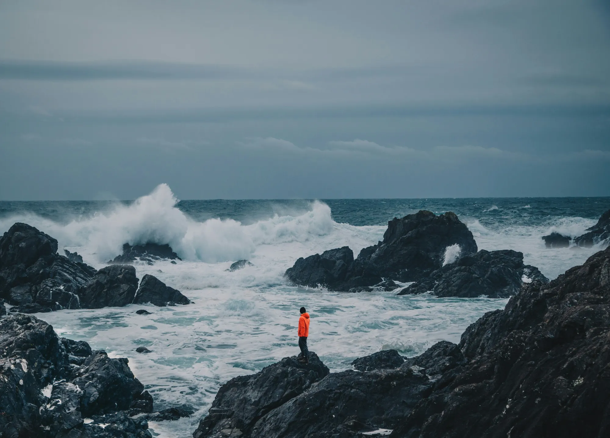 Person in orange jacket standing on rocks beside crashing waves