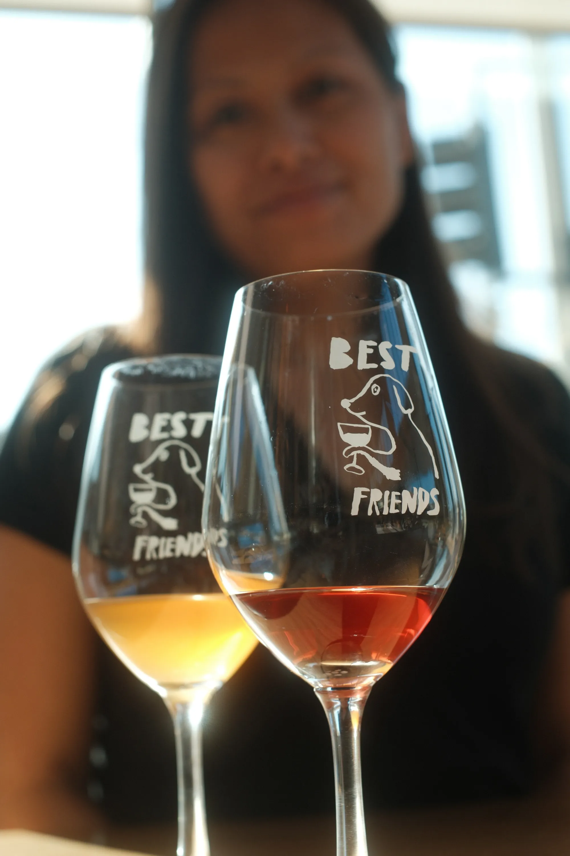 Two engraved "Best Friends" wine glasses on a wooden table with a person soft-focused behind them